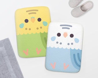 May include: Two bath mats featuring cartoon budgies. One mat is yellow and green with a light pink heart on the bottom. The other mat is white and blue with a light pink heart on the bottom.