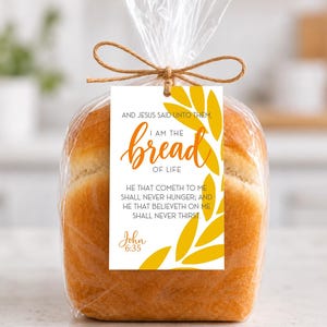 May include: A loaf of golden-brown bread packaged in a clear plastic bag tied with a brown twine bow. A white rectangular tag features yellow leaf graphics and the text: AND JESUS SAID UNTO THEM, I AM THE BREAD OF LIFE, HE THAT COMETH TO ME SHALL NEVER HUNGER; AND HE THAT BELIEVETH ON ME SHALL NEVER THIRST. John 6:35.
