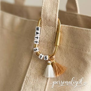 May include: A gold-toned carabiner clip attached to a beige canvas tote bag. The clip features white square beads with black lettering spelling MATT 5 16, accented by two small hanging tassels in white and tan colors, dangling from the bottom loop.