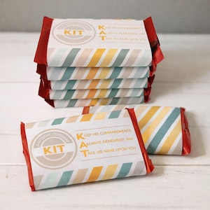 May include: A stack of wrapped rectangular items with red edges. The wrappers have a white background with diagonal stripes in shades of blue, yellow, and orange. Text on the wrapper reads "Baptismal Covenant Kit" and "Keep His Commandments, Always Remember Him, Take His Name Upon You."