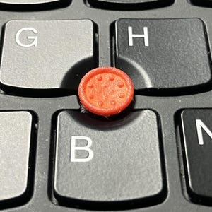 Puede incluir: Una tapa de puntero circular de color naranja brillante con una superficie texturizada que presenta pequeños puntos en relieve. La tapa está colocada centralmente entre las teclas G, H y B en un teclado de computadora negro con letras blancas.