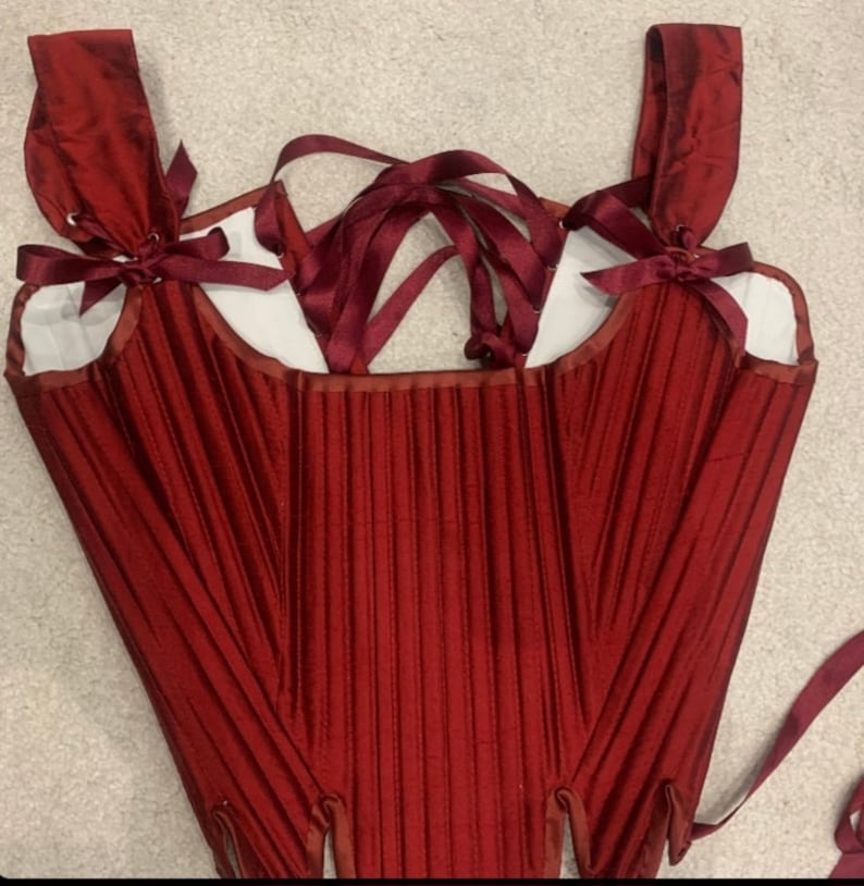 May include: A red satin corset with boning and a lace-up back. The corset has adjustable straps and a bow at the top of each strap.