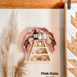 May include: A pink and white wooden shelf with multiple levels, holding various items including jars, crystals, and a candle. The shelf is mounted on a white wall, with text that reads "Pink Stain {top}".