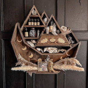 May include: A dark walnut stained wooden shelf shaped like a mountain range with gold moons and ferns. The shelf has multiple compartments and is decorated with crystals, a skull, and a small picture frame. The text "Dark Walnut Stain W/gold moons and ferns + cosmic moth drawer knob add on" is visible at the bottom of the image.