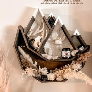 May include: A dark walnut stained mountain-shaped shelf with white snowy peaks. The shelf is decorated with antlers, a skull, crystals, dried flowers, and small jars. The text on the shelf reads "Dark Walnut Stain w/ Solid Snowy Peaks & No Moon Phases."