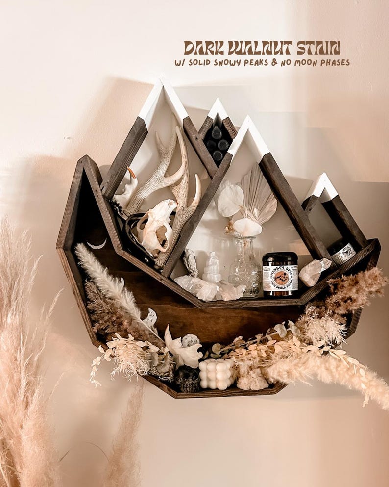 May include: A dark walnut stained mountain-shaped shelf with white snowy peaks. The shelf is decorated with antlers, a skull, crystals, dried flowers, and small jars. The text on the shelf reads "Dark Walnut Stain w/ Solid Snowy Peaks & No Moon Phases."