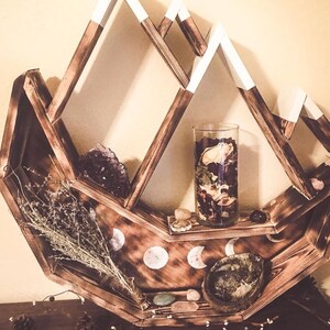 May include: A wooden mountain-shaped shelf with white-tipped peaks. The shelf is stained brown and holds a glass vase with dried flowers, crystals, and decorative stones. The shelf also features moon phase designs.