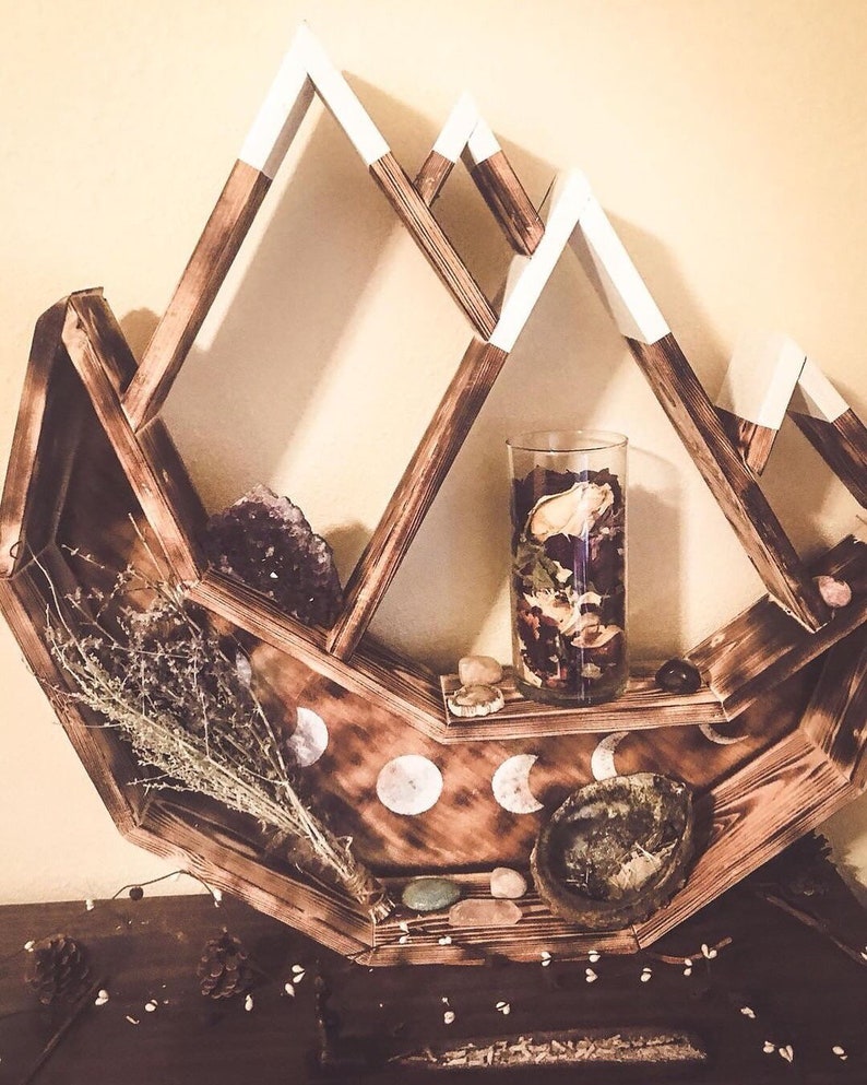May include: A wooden mountain-shaped shelf with white-tipped peaks. The shelf is stained brown and holds a glass vase with dried flowers, crystals, and decorative stones. The shelf also features moon phase designs.