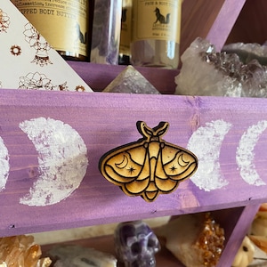May include: A purple wooden shelf with two tiers, featuring a wooden moth-shaped drawer pull. The shelf is decorated with white painted moons and holds various crystals, including amethyst, clear quartz, and citrine. The top tier also features two glass bottles with labels that read "Coffee & Peppermint Whipped Body Butter" and "Lavender Face & Body Toner".