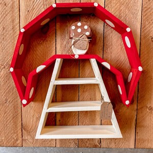 May include: A whimsical, mushroom-shaped shelf with a red, polka-dotted top and a white, three-tiered shelf. A small, mushroom-shaped ornament is centered above the shelf. The shelf is mounted on a wooden background.