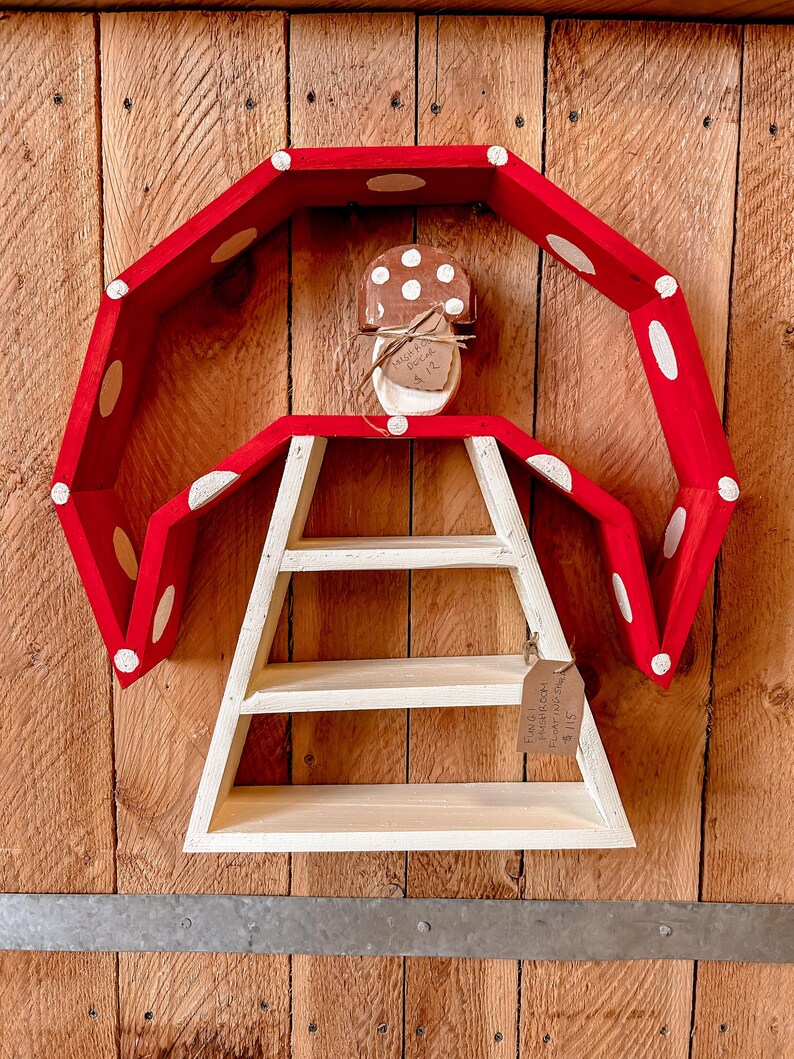 May include: A whimsical, mushroom-shaped shelf with a red, polka-dotted top and a white, three-tiered shelf. A small, mushroom-shaped ornament is centered above the shelf. The shelf is mounted on a wooden background.
