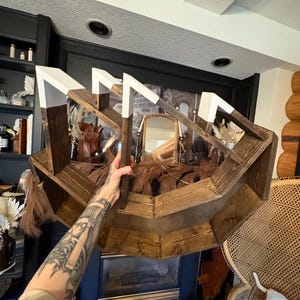 May include: A rustic, wooden shelf with a unique geometric design. The shelf is stained a warm brown with white accents on the top edges. It is filled with decorative items, including dried plants and a small mirror.