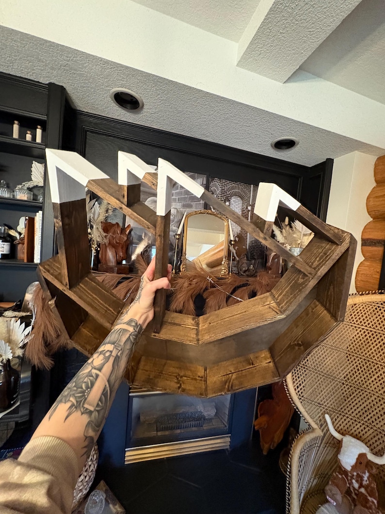 May include: A rustic, wooden shelf with a unique geometric design. The shelf is stained a warm brown with white accents on the top edges. It is filled with decorative items, including dried plants and a small mirror.