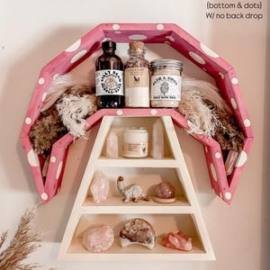 May include: A pink and white polka-dotted shelf with three levels, displaying various items. The top shelf holds bottles and jars, while the lower shelves showcase crystals and decorative objects. The shelf is mounted on a white wall.