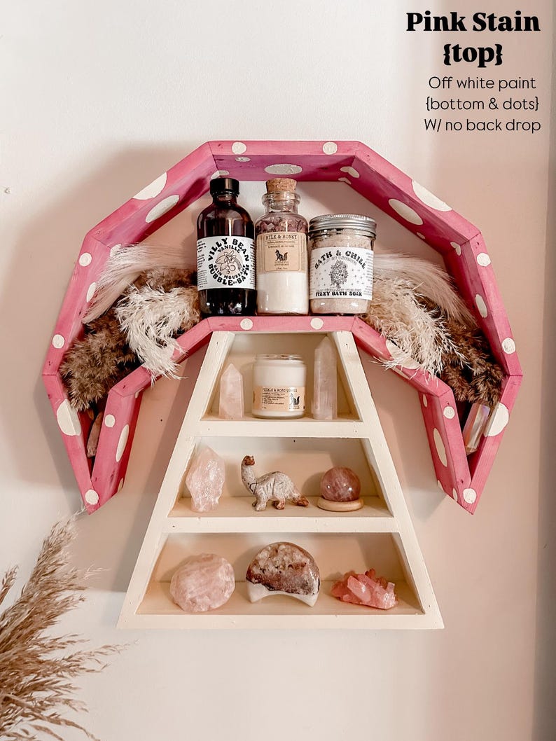 May include: A pink and white polka-dotted shelf with three levels, displaying various items. The top shelf holds bottles and jars, while the lower shelves showcase crystals and decorative objects. The shelf is mounted on a white wall.