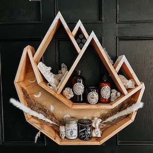 May include: Wooden shelf in a mountain shape, stained golden oak. The shelf holds various items, including crystals, jars, and bottles with labels. The shelf is decorated with dried plants and moon phases.