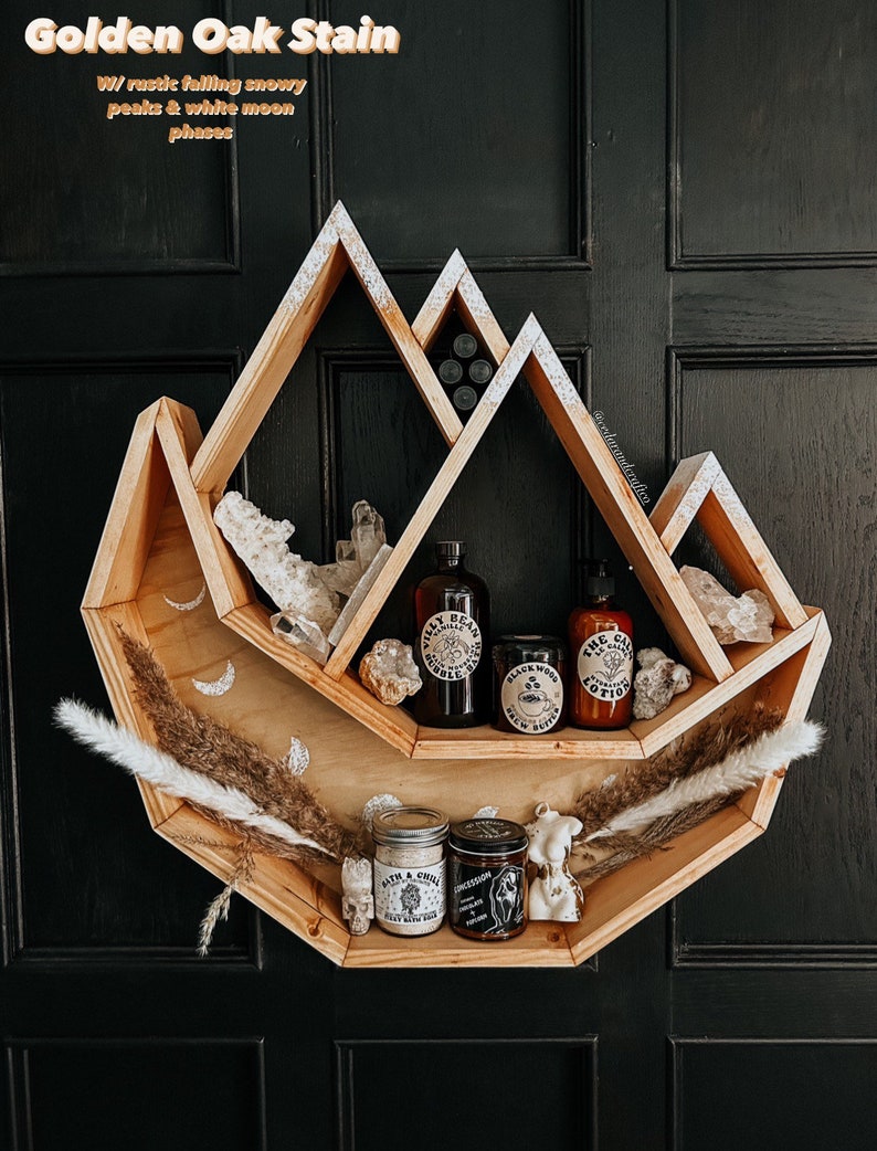 May include: Wooden shelf in a mountain shape, stained golden oak. The shelf holds various items, including crystals, jars, and bottles with labels. The shelf is decorated with dried plants and moon phases.