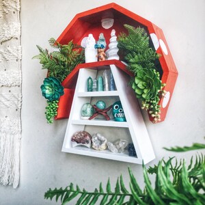 May include: A red and white wooden shelf in a unique geometric shape, adorned with crystals, decorative figurines, and faux greenery. The shelf is mounted on a white wall, creating a visually appealing display for home decor.