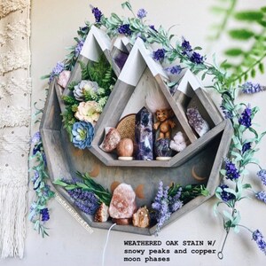 May include: A weathered oak stain wooden shelf in a mountain shape with white snowy peaks and copper moon phases. The shelf is decorated with crystals, a troll figurine, and artificial flowers. The shelf is surrounded by purple floral garland.