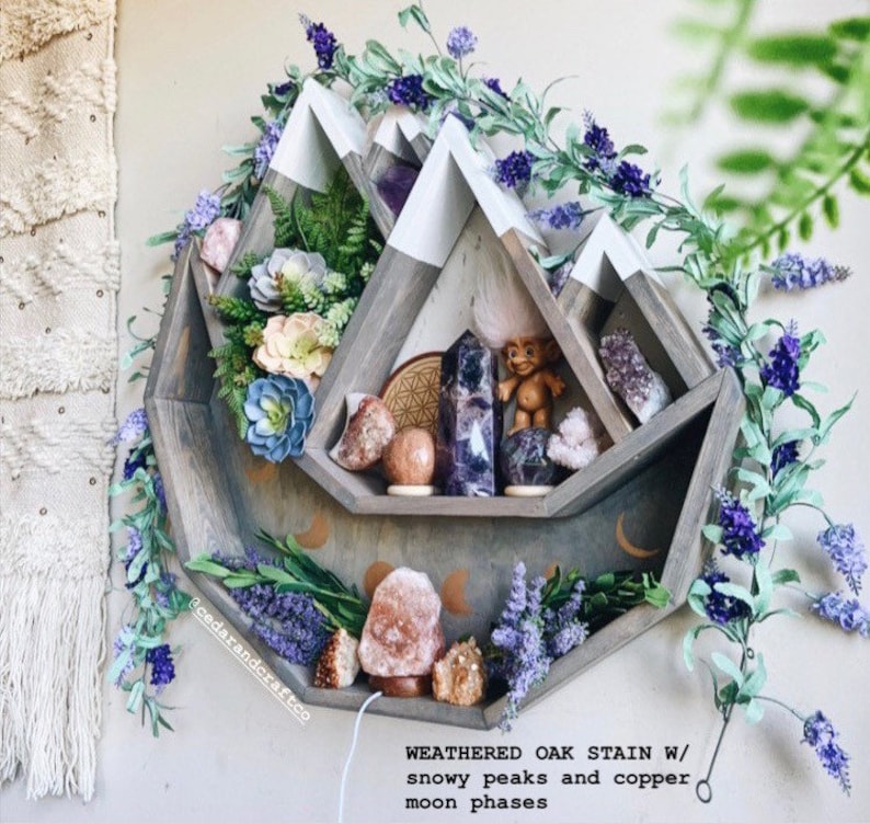May include: A weathered oak stain wooden shelf in a mountain shape with white snowy peaks and copper moon phases. The shelf is decorated with crystals, a troll figurine, and artificial flowers. The shelf is surrounded by purple floral garland.
