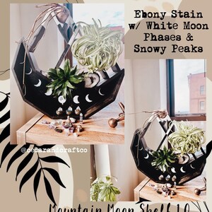 May include: Ebony-stained wooden shelf with white moon phases and snowy peaks design. The shelf holds various plants and crystals. The text on the shelf reads "Mountain Moon Shelf 1.0".