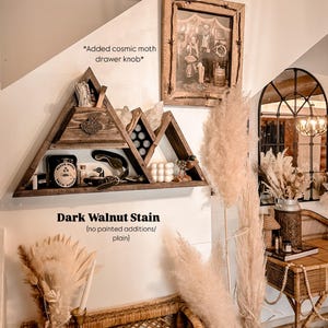 May include: A rustic wooden shelf in a mountain shape, stained dark walnut, displays decorative items. A framed black and white photo hangs above. The text "Added cosmic moth drawer knob" is visible. A white rotary phone sits below.