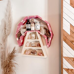 May include: A unique, pink polka-dotted shelf with a wooden A-frame design. The shelf displays various items, including small jars, bottles, crystals, and decorative objects. The shelf is mounted on a white wall, with a wooden accent on the right.