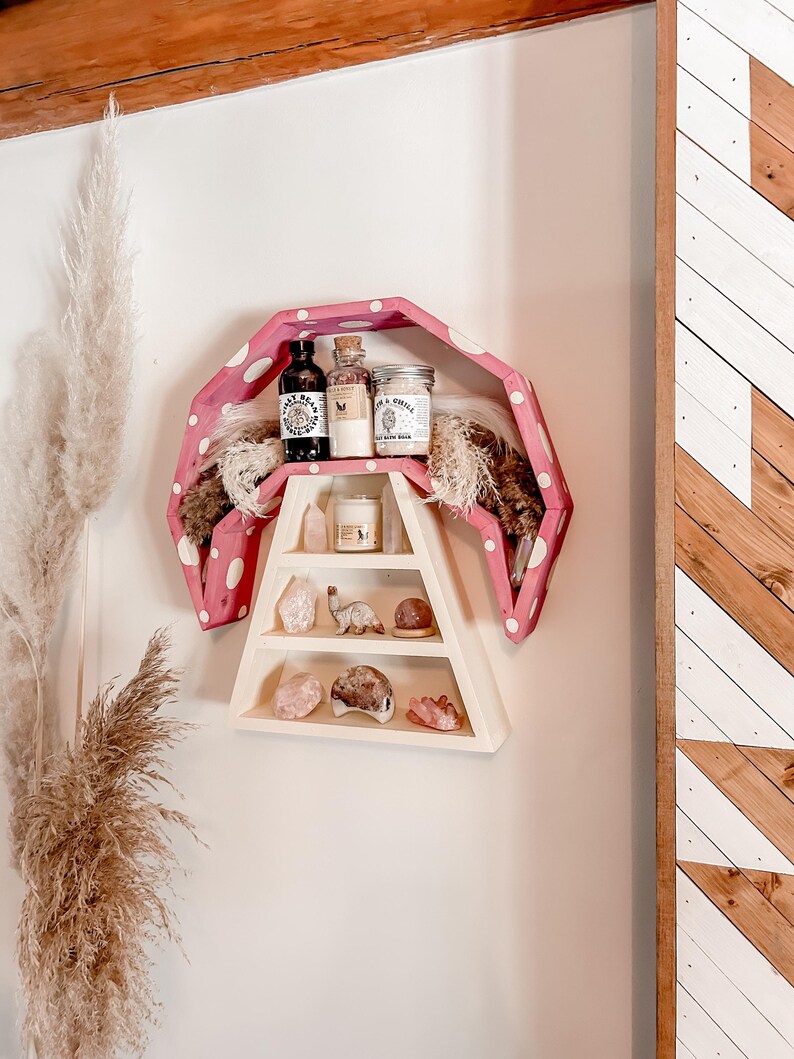 May include: A unique, pink polka-dotted shelf with a wooden A-frame design. The shelf displays various items, including small jars, bottles, crystals, and decorative objects. The shelf is mounted on a white wall, with a wooden accent on the right.