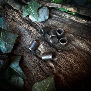 Könnte beinhalten: Eine Sammlung kleiner, zylindrischer Metallperlen mit dunkler, gealterter Oberfläche. Die Perlen liegen auf einer verwitterten, dunkelbraunen Holzoberfläche, umgeben von grünen Efeublättern und einem kleinen grauen Stein. Die Perlen haben eine hohle Mitte.