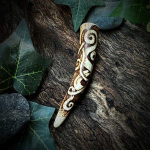 Puede incluir: Colgante de cuerno tallado con un diseño de espirales de inspiración celta. El colgante es de color crema claro con detalles grabados en marrón oscuro. Mide aproximadamente 13 cm de largo y se muestra sobre madera envejecida.