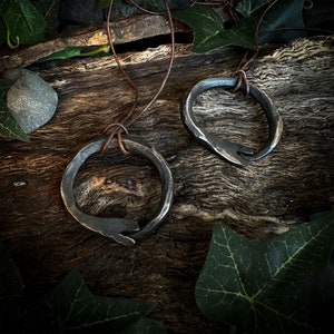 May include: Two handcrafted, dark metal pendant necklaces. Each pendant is a circular design with a unique, open-ended shape, suspended from a brown leather cord. The pendants rest on a rustic wooden surface, accented with green foliage.