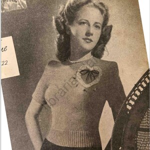 May include: A black and white photo of a woman wearing a knitted sweater with a heart-shaped design. The sweater has a fitted silhouette and short sleeves. The woman is wearing a pearl necklace and has her hair styled in a soft wave.