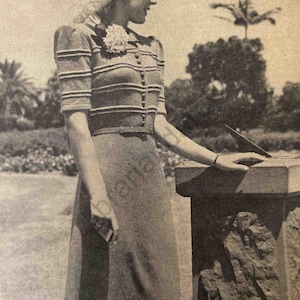 May include: Vintage knitted two-piece outfit featuring a button-down top with horizontal stripes and a matching skirt. The ensemble includes a hat and a floral accent. The outfit is worn outdoors near a stone structure.