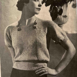 May include: A black and white photograph of a woman wearing a knitted jumper with a round neckline and short sleeves. The jumper has a textured pattern and is decorated with buttons. The text "LADY'S JUMPER "DOREEN" DESIGN" is printed at the bottom of the image.