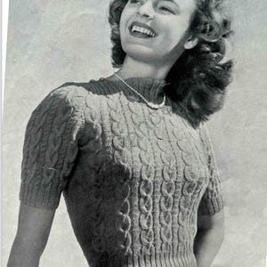 Puede incluir: Primer plano de una mujer con un suéter gris de punto de cable con mangas cortas. El suéter tiene un cuello y un dobladillo de canalé. La mujer sonríe y mira hacia un lado.