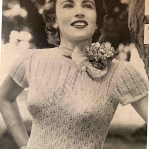 Puede incluir: Una mujer lleva un suéter de punto blanco con un delicado patrón de encaje. Lleva una flor en el hombro y tiene una bufanda atada alrededor del cuello.