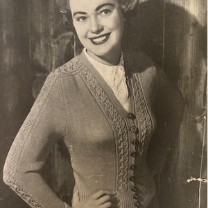 May include: A vintage black and white image of a woman wearing a light-colored knitted cardigan with a V-neck and button closure. The cardigan has decorative patterns along the edges and sleeves. The woman is smiling.