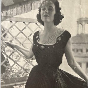Puede incluir: Un vestido negro sin mangas con un ribete de encaje blanco y un diseño en forma de estrella. El vestido lo lleva una mujer con el pelo oscuro y una expresión neutral. La mujer está de pie frente a una valla de enrejado.