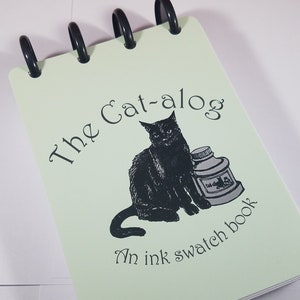May include: A green spiral bound notebook with a black cat illustration and the text "The Cat-alog An ink swatch book".