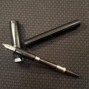 May include: A black fountain pen with a silver nib and accents, disassembled on a patterned surface. The pen's cap and barrel are separate, showcasing its components. The pen is designed for writing.