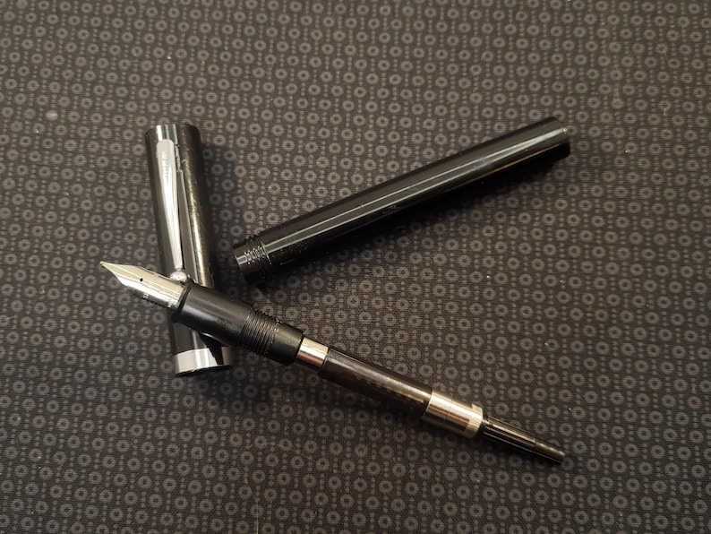 May include: A black fountain pen with a silver nib and accents, disassembled on a patterned surface. The pen's cap and barrel are separate, showcasing its components. The pen is designed for writing.