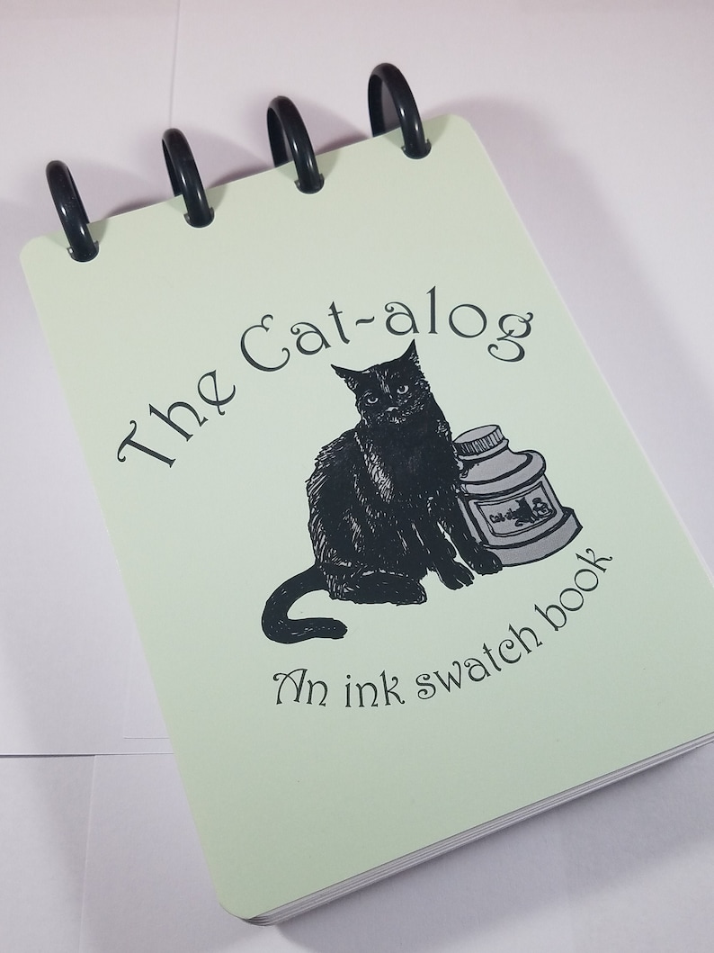 May include: A green spiral bound notebook with a black cat illustration and the text "The Cat-alog An ink swatch book".