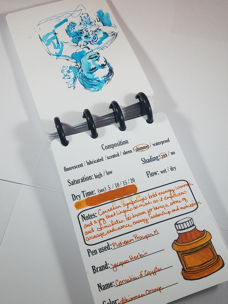 May include: A close-up of a handwritten ink swatch card with notes about the ink's properties. The ink is a vibrant orange color with a shimmer finish. The card includes the brand name "Jacques Herbin" and the ink name "Cornaline d'Egypte".