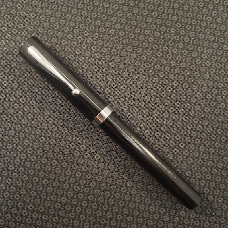 May include: A black fountain pen with silver accents. The pen has a shiny, smooth finish and a silver clip. The pen's design is simple and elegant, suitable for writing or calligraphy. The pen is set against a dark patterned background.