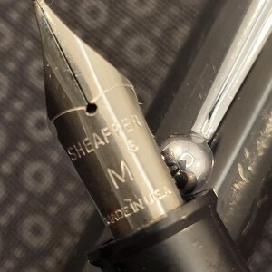 May include: Close-up of a silver fountain pen nib with the brand name "SHEAFFER" and the letter "M" engraved on it. The pen is black and silver, with "MADE IN USA" printed on the nib.