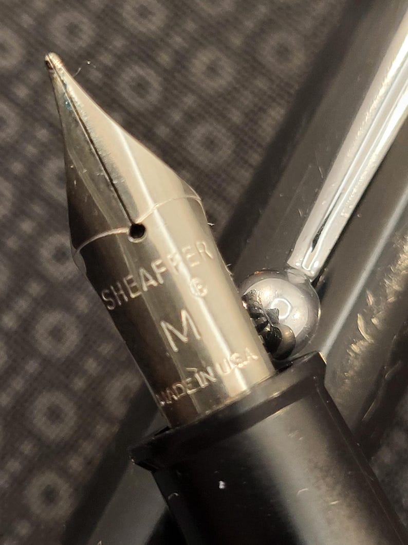 May include: Close-up of a silver fountain pen nib with the brand name "SHEAFFER" and the letter "M" engraved on it. The pen is black and silver, with "MADE IN USA" printed on the nib.