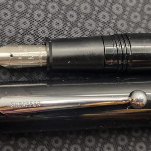 May include: Close-up of a black fountain pen with a silver nib and accents. The nib is marked "SHEAFFER" and "M". The pen's cap is also black with a silver clip that reads "SHEAFFER". The pen is a classic writing instrument.