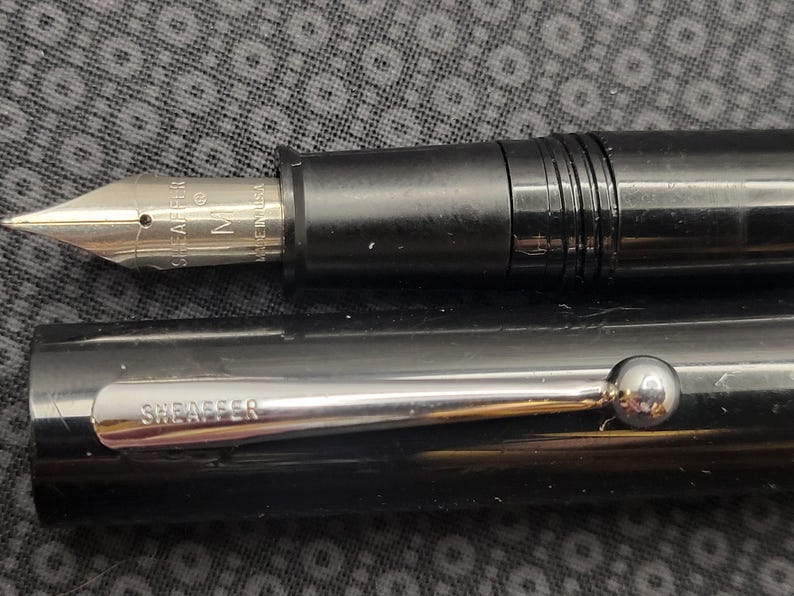 May include: Close-up of a black fountain pen with a silver nib and accents. The nib is marked "SHEAFFER" and "M". The pen's cap is also black with a silver clip that reads "SHEAFFER". The pen is a classic writing instrument.
