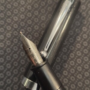 May include: Close-up of a black fountain pen with a silver nib and accents. The pen's nib is marked "SHEAFFER" and "M MADE IN USA". The pen's cap is open, revealing the nib and part of the ink reservoir.
