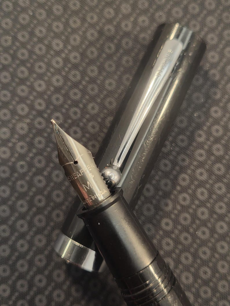 May include: Close-up of a black fountain pen with a silver nib and accents. The pen's nib is marked "SHEAFFER" and "M MADE IN USA". The pen's cap is open, revealing the nib and part of the ink reservoir.